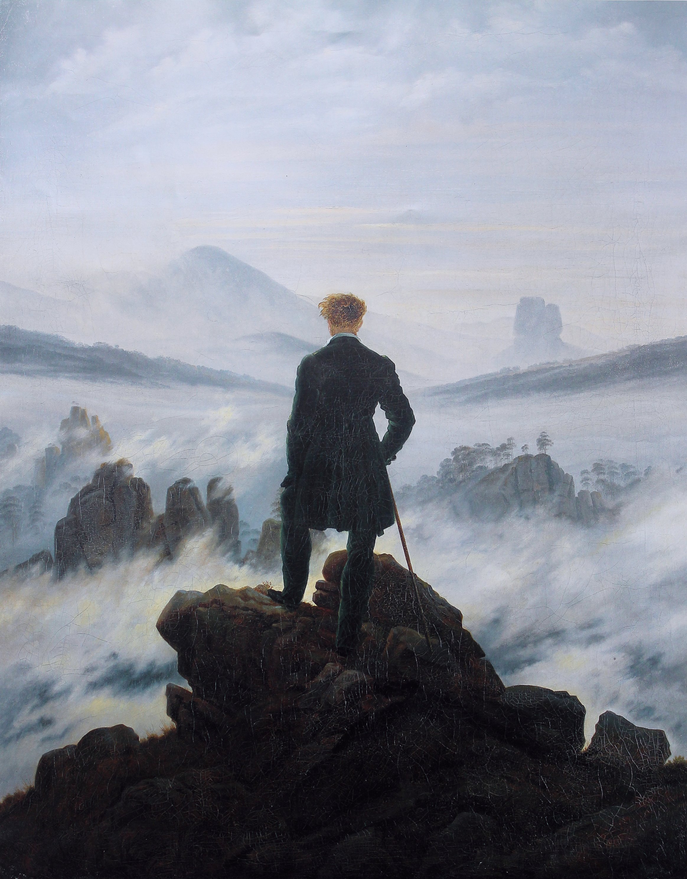 A man in a dark coat stands on a rocky precipice, back to the viewer, gazing over an ocean of fog with mountain peaks emerging
